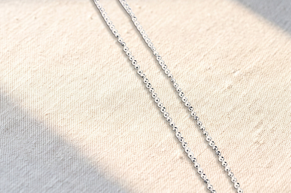 Srebrny łańcuszek kulkowy z diamentowanymi kulkami o grubości 1,2 mm, złożony w dwie równoległe nitki, leży ukośnie na lnianym obrusie w plamie porannego słońca; przy kulkach drobne cienie