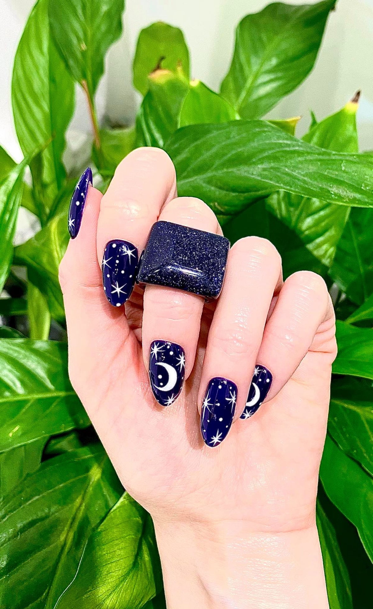 Pierścionek Noc Kairu prezentowany na dłoni modelki z kosmicznym manicure na tle zielonych roślin.
