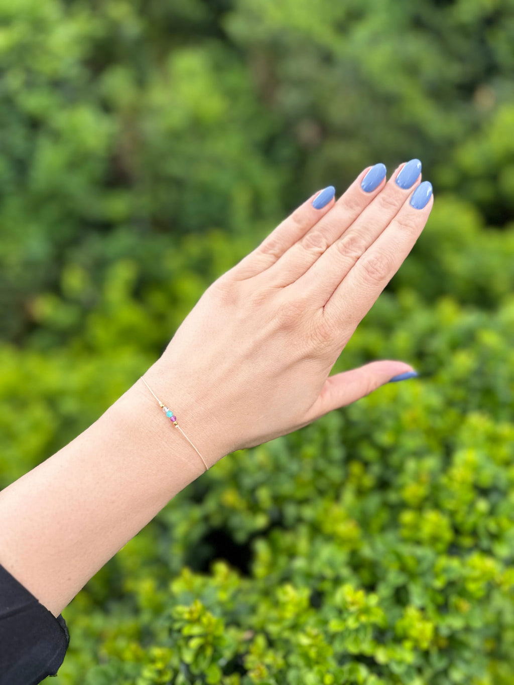 Bransoletka z minerałów na dłoni z niebieskim manicure na tle zielonych roślin, marka Brazi