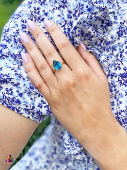 Złoty pierścionek z topazem London Blue o szlifie kropli na tle kwiecistego materiału, biżuteria Brazi.