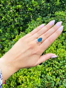 Pierścionek z fasetowanym topazem London Blue w złotej oprawie na tle zielonego żywopłotu, biżuteria Brazi.